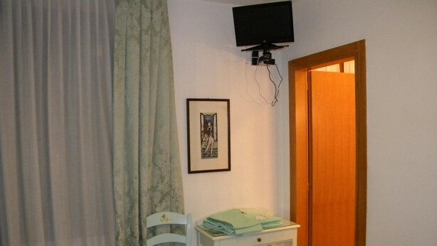 Una habitación pequeña con una silla blanca, una mesa con ropa doblada, un cuadro en la pared y un televisor montado encima de la mesa.