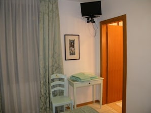 A small room with a white chair, a table with folded clothes, a framed picture on the wall, and a television mounted above the table. - Adua Hotel (Venice)