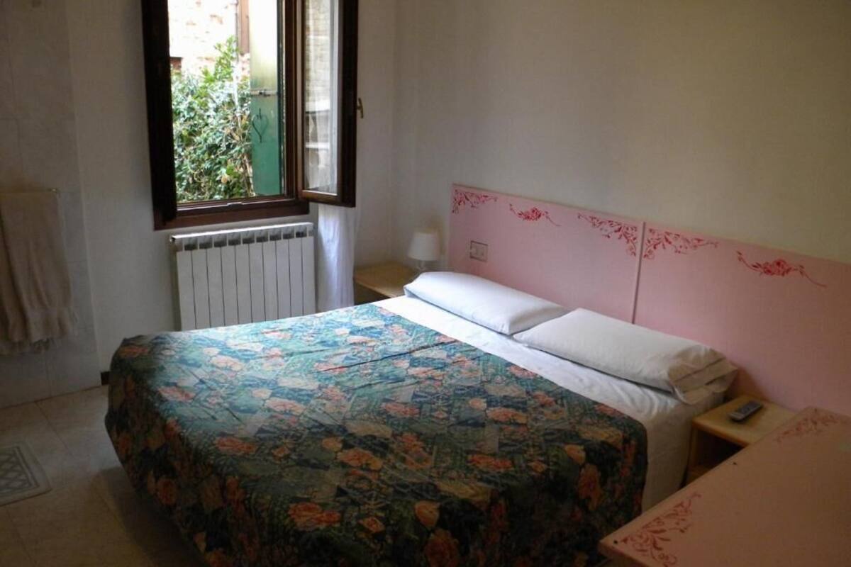 A bedroom with a floral bedspread, a pink headboard, a bedside table with a lamp, and a window with a view of greenery.
