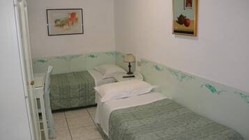 A small, simple room with two single beds, a chair, a table with a lamp, and a framed picture on the wall.