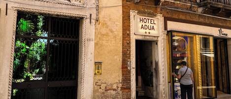 Un edificio histórico con una entrada arqueada destacada, un cartel que dice 'HOTEL ADUA' y un escaparate de vidrio moderno.