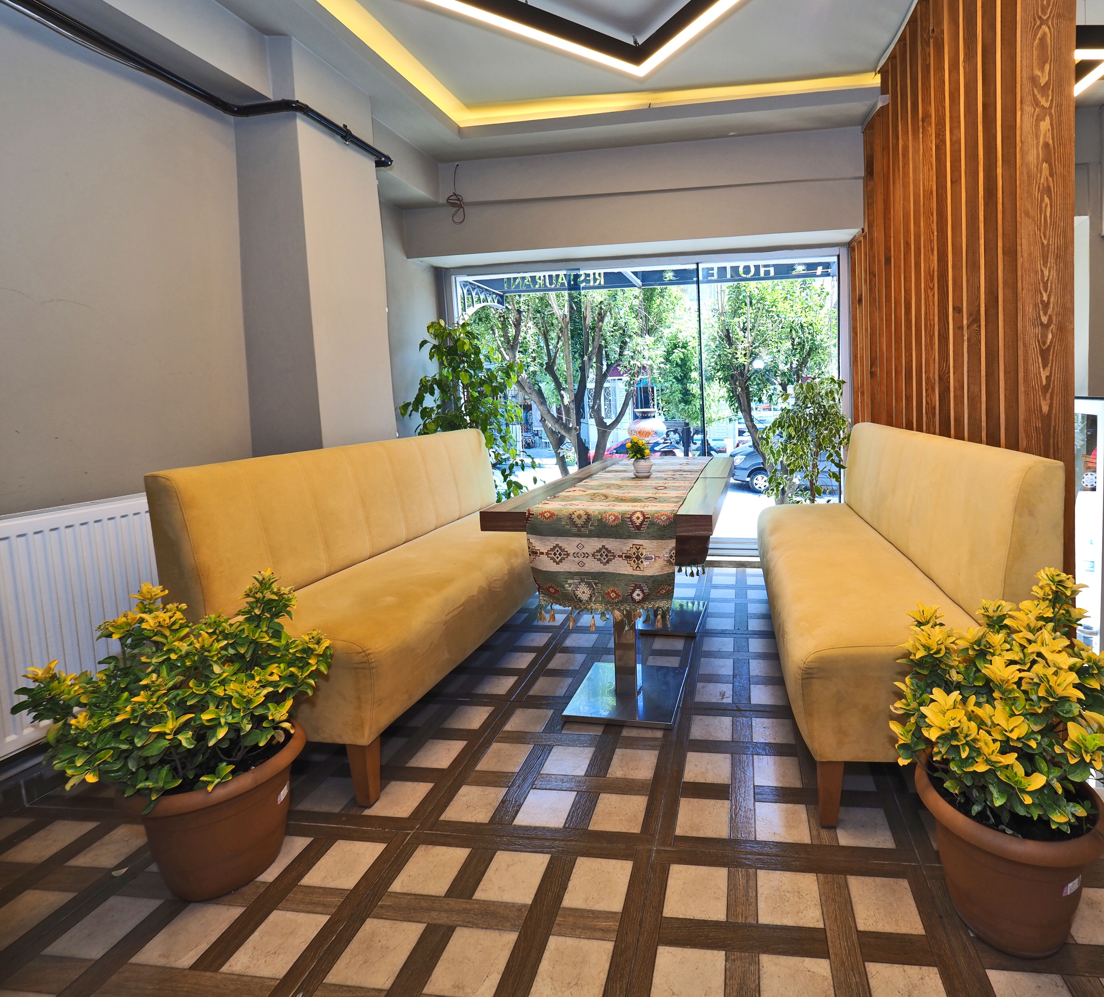 Ein modernes Interieur mit zwei Sofas, die einem gemusterten Tisch gegenüberliegen, Zimmerpflanzen und Blick auf die Straße durch große Fenster.