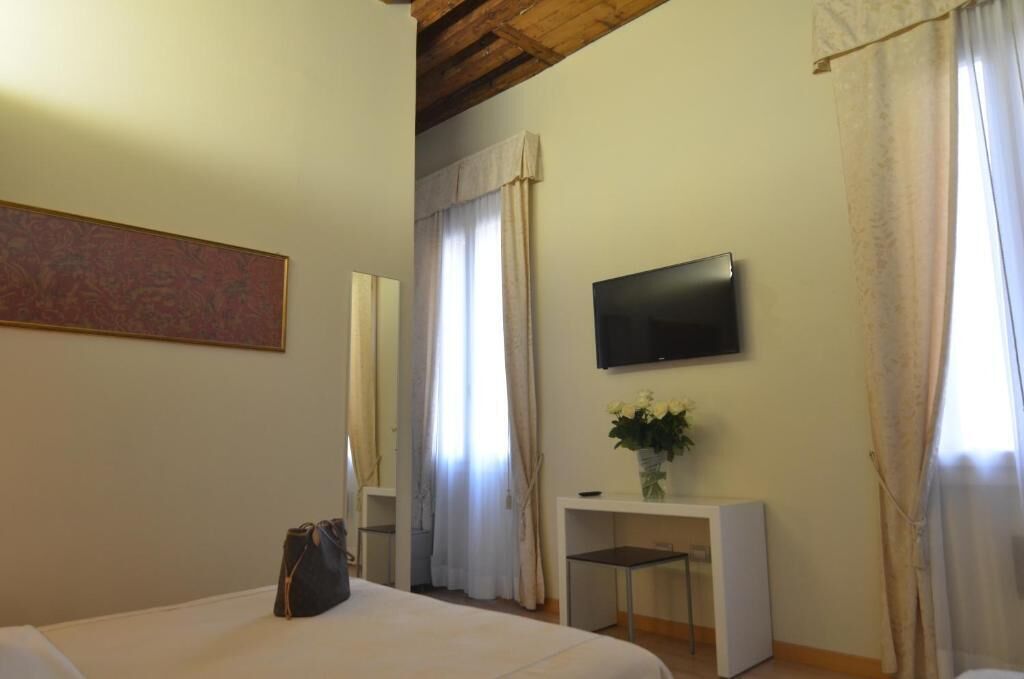 Una camera d'albergo con un letto, una scrivania con un vaso di fiori, una TV a schermo piatto montata a parete e una finestra con tende.