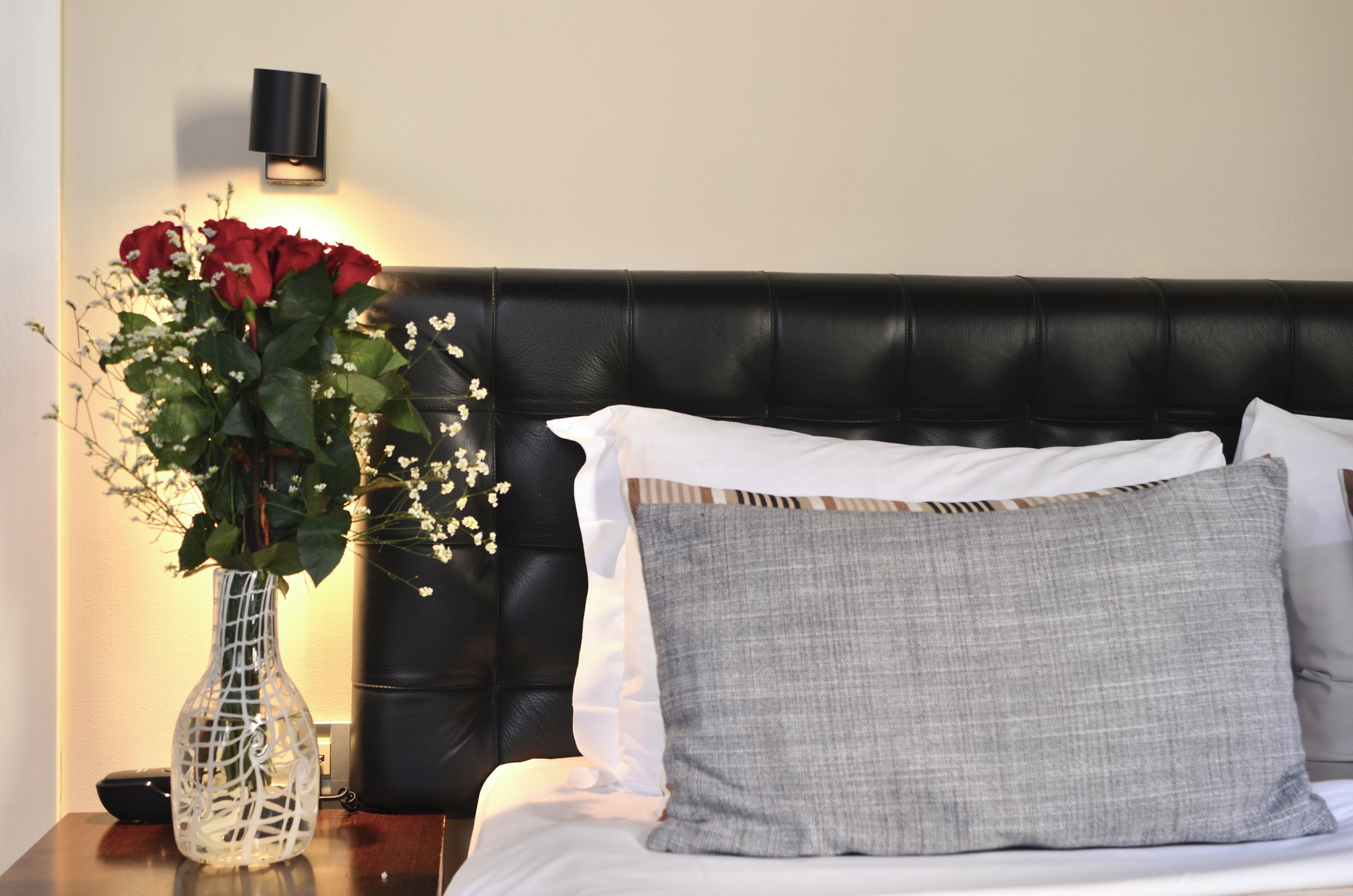 A bed with a black headboard, white and grey pillows, and a white vase with red flowers on a bedside table.