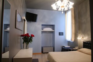 Habitación de hotel con cama, mesita de noche, espejo, televisor montado en la pared, armario y ventana con cortinas.