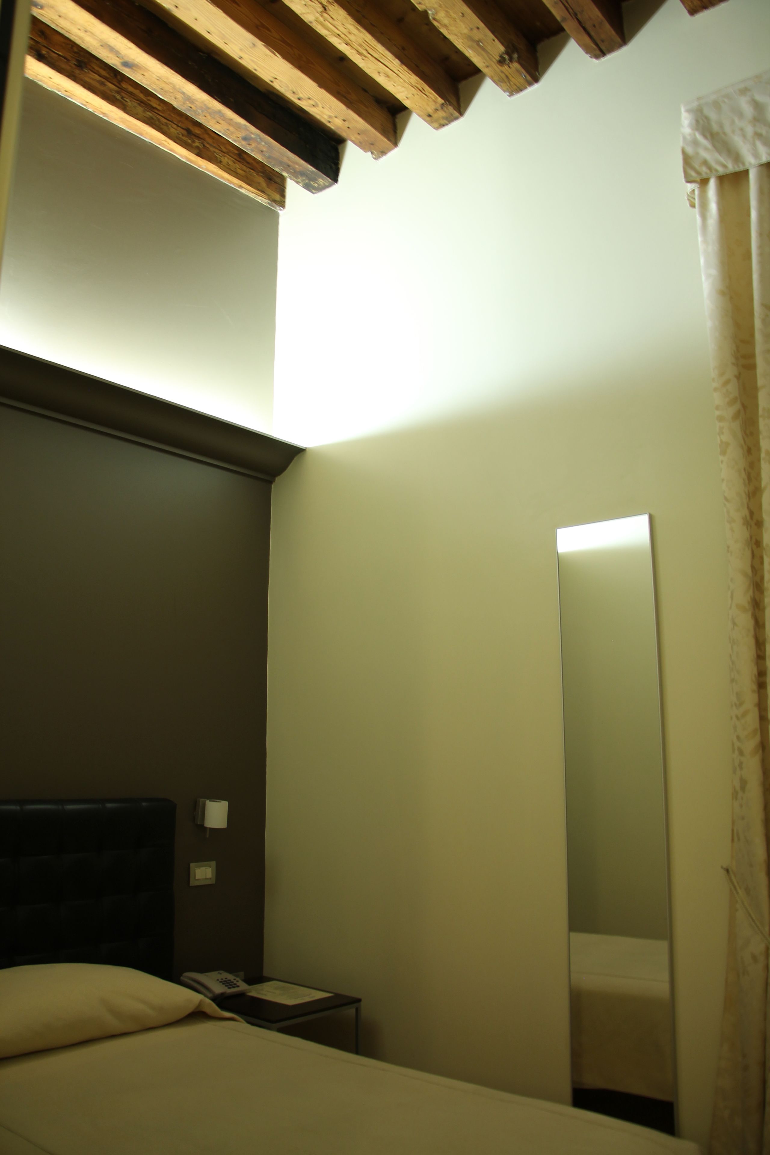A hotel room with a bed, a nightstand with a phone, a mirror, and a wooden ceiling beam.
