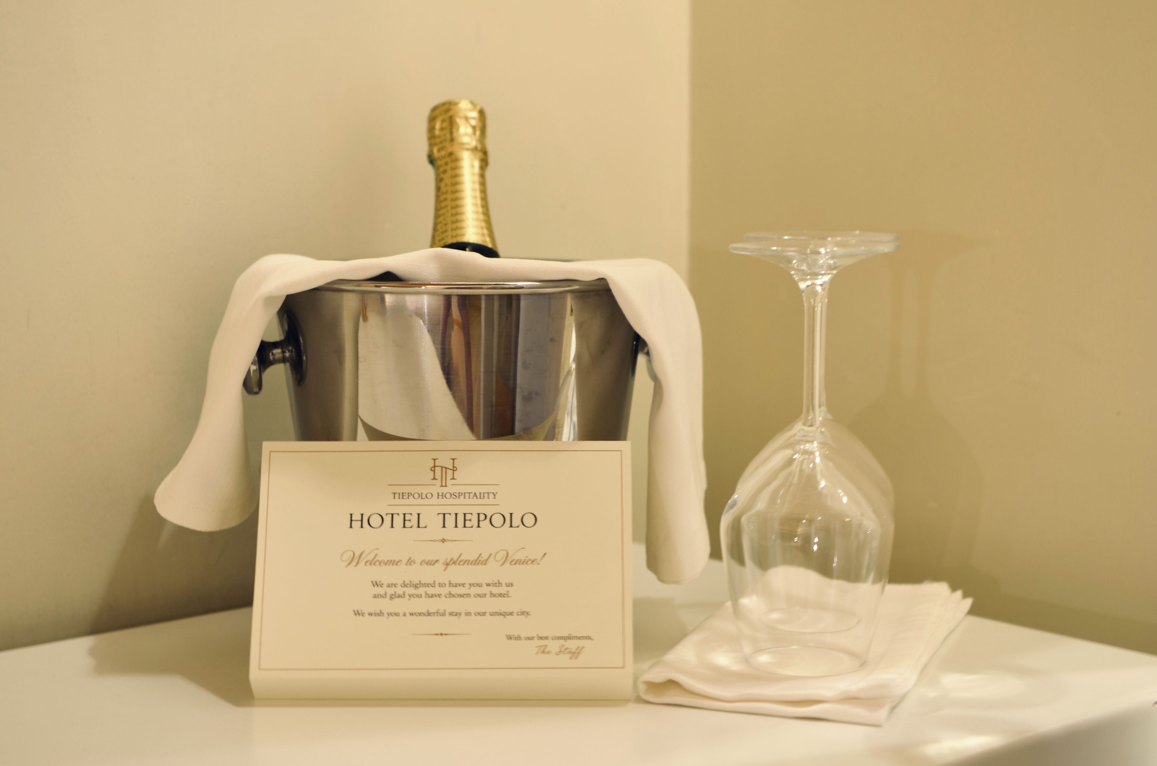 A bottle of champagne in a metal bucket, a glass, and a welcome card for HOTEL TIEPOLO.