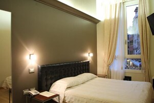 A hotel room with a dark headboard, a bedside telephone, and a window with curtains.