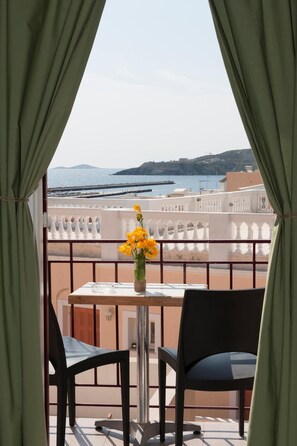 A balcony with a table and chairs, a vase with yellow flowers, and a view of the sea and hills. - Paradise Rooms (Syros)