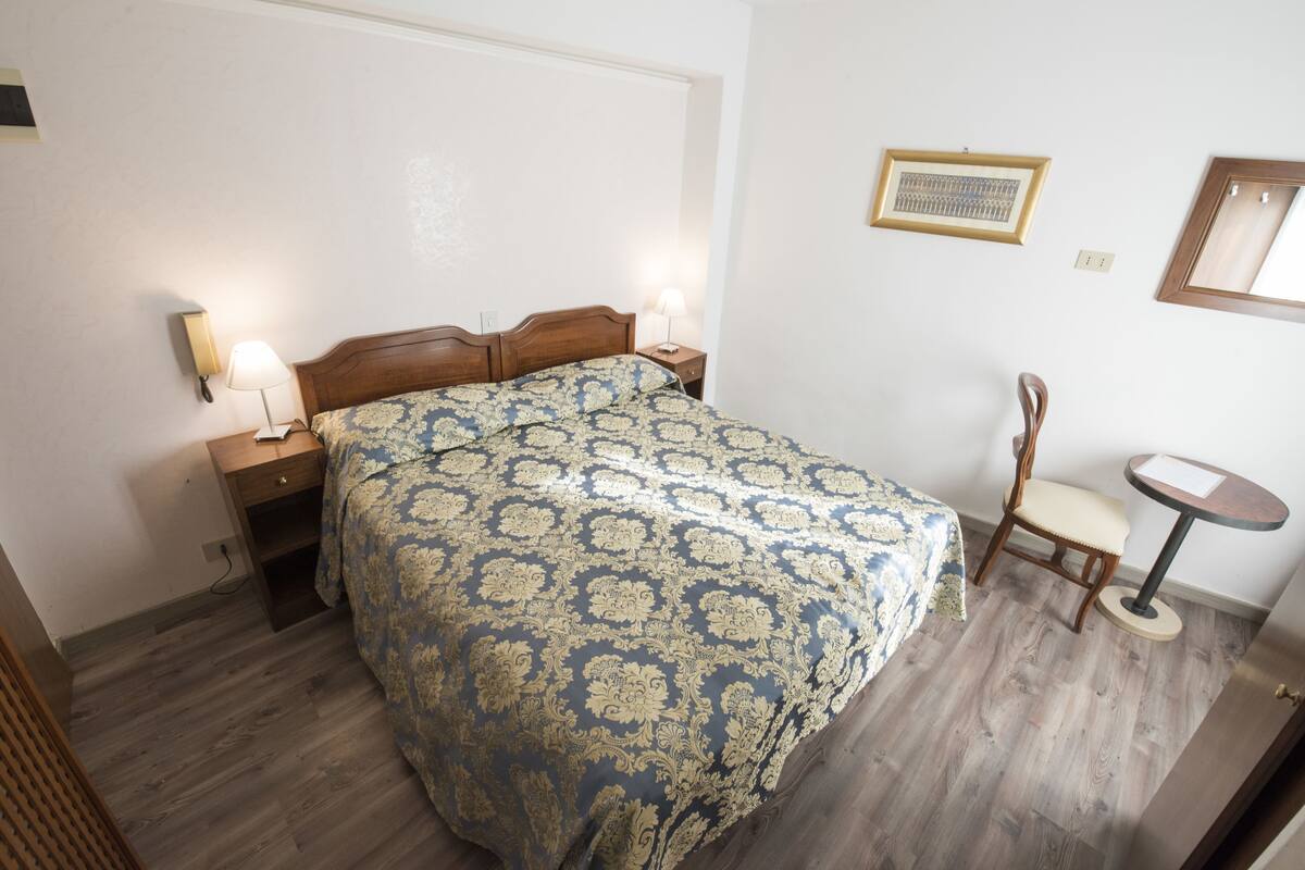 A bedroom with a wooden bed, two bedside tables with lamps, a chair, a small round table, and a framed picture on the wall.