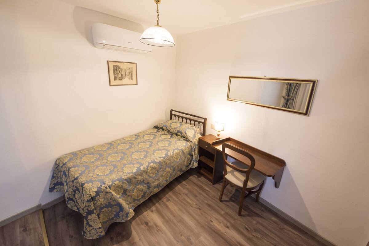 A bedroom with a bed, a desk with a chair, a mirror, and an air conditioning unit.