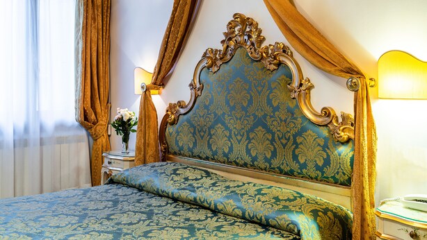 A bed with a blue and gold headboard, a bedside table with a lamp, and a window with curtains. - Hotel Al Vagon (Venice)
