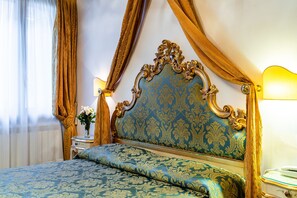 Una cama con cabecera azul y dorada, una mesita de noche con lámpara y una ventana con cortinas.