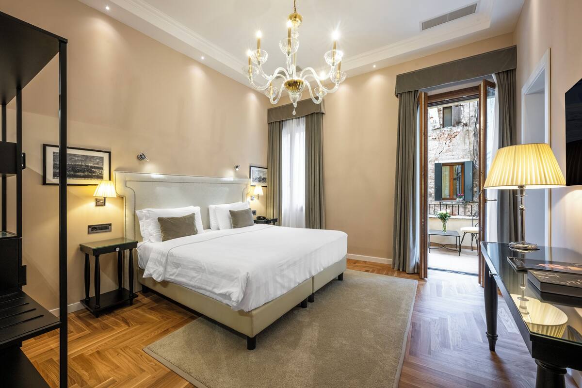 A hotel room with a large bed, a chandelier, a desk with a lamp, and a view of a courtyard through a window.