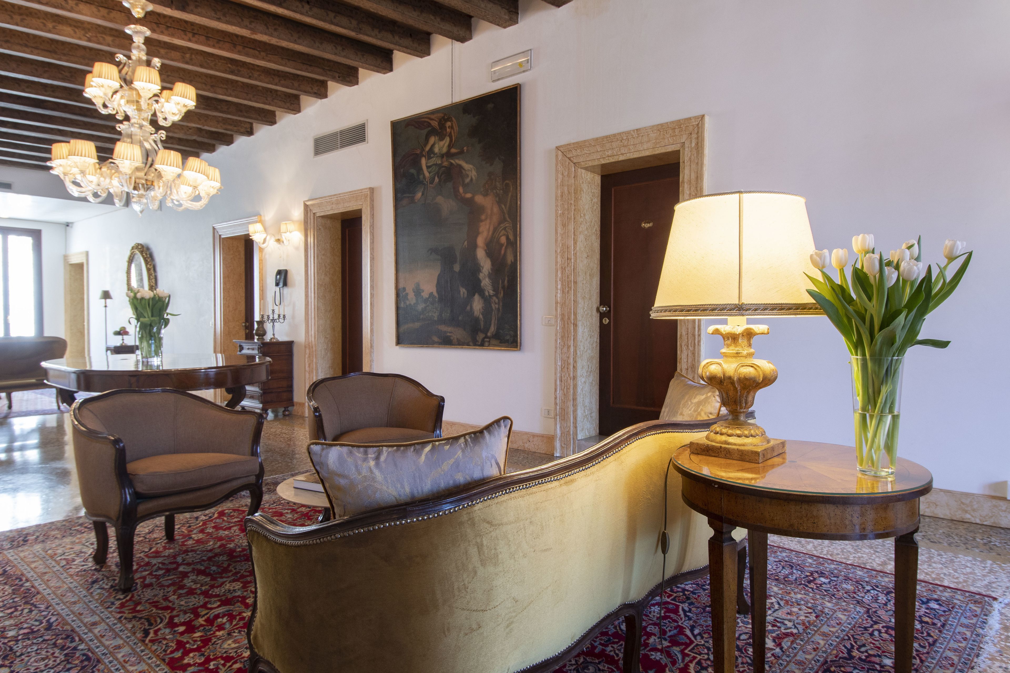 A lobby with a grand chandelier, a large painting, and a table with a lamp and vase of flowers.