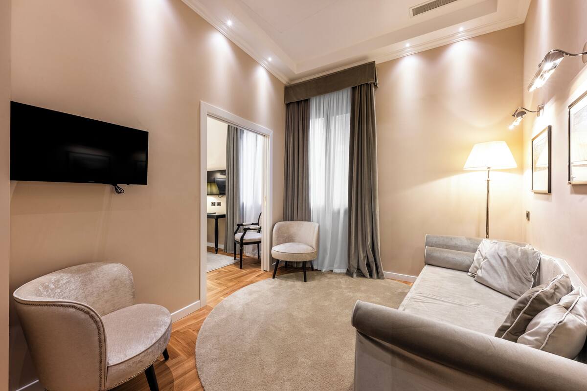 A modern hotel room with a flat-screen TV mounted on the wall, a round beige carpet, a grey sofa, a white armchair, and a dining area visible through a doorway.