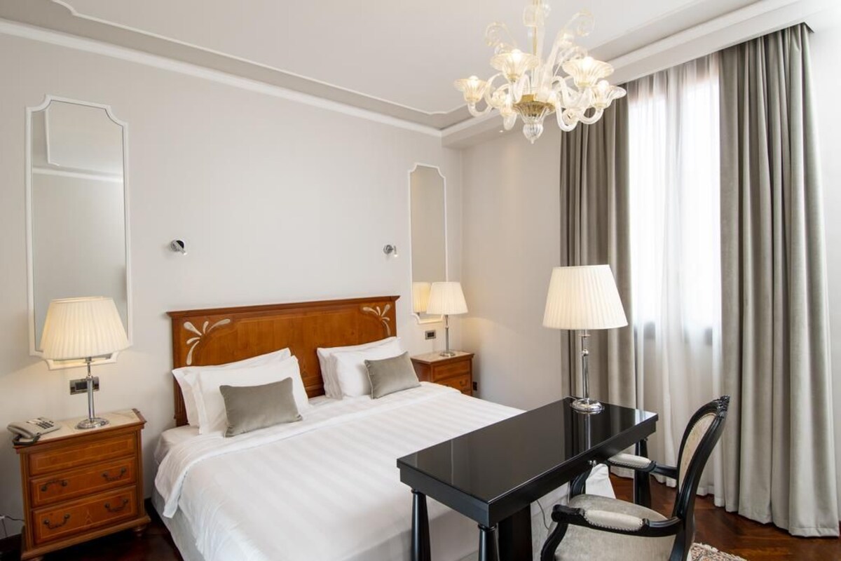 Hotelkamer met een groot bed, twee nachtkastjes met lampen, een houten hoofdeinde, een zwarte tafel met een stoel, en een kroonluchter.