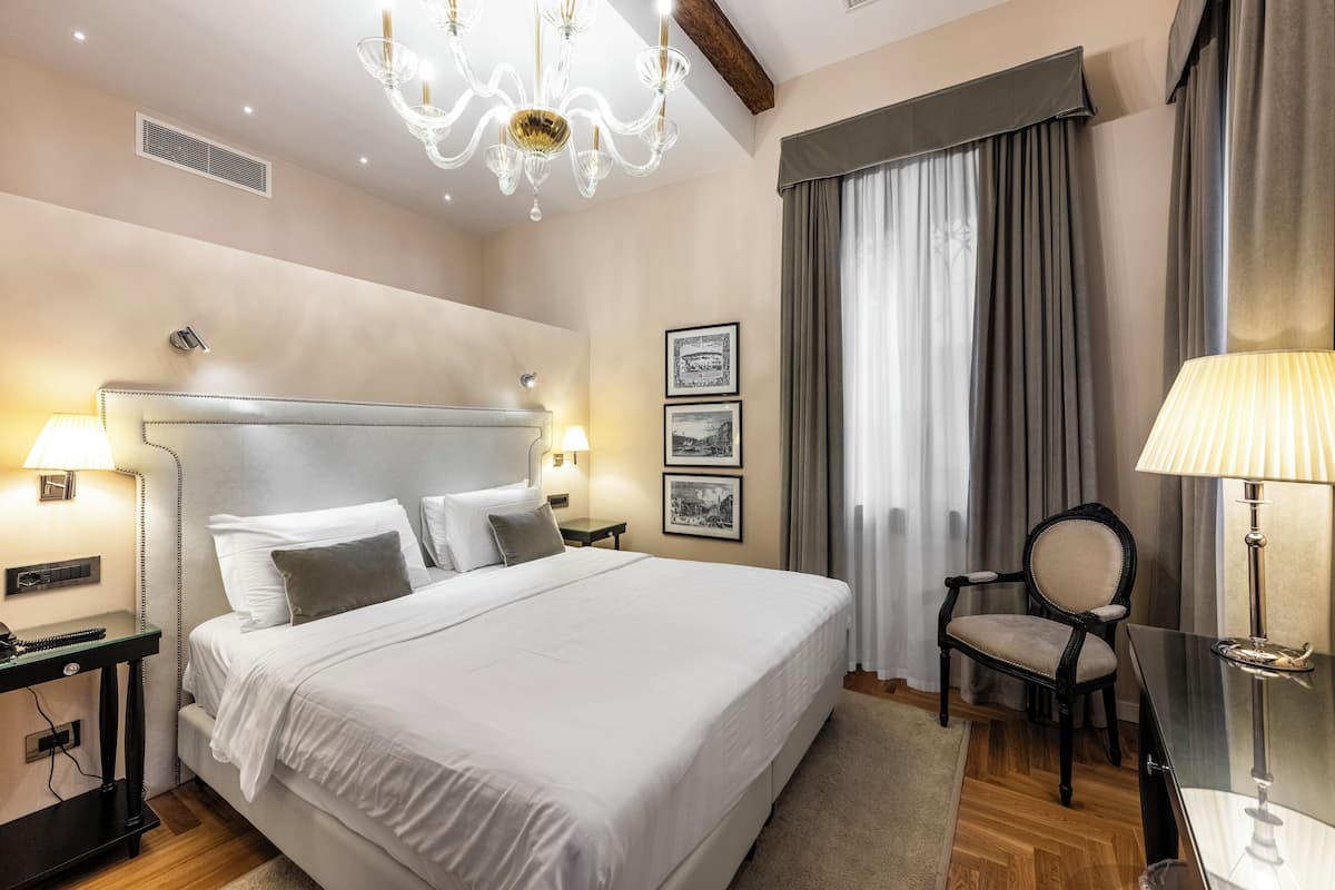 A hotel room with a large bed, two bedside lamps, a chair, a desk, and a chandelier.