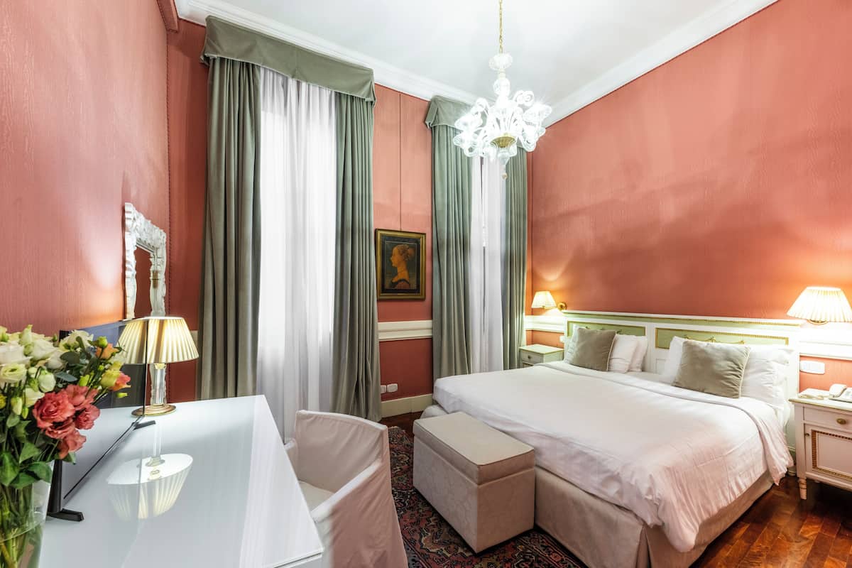 A hotel room with a large bed, a desk with a lamp, a chair, a small table with flowers, and a painting on the wall.