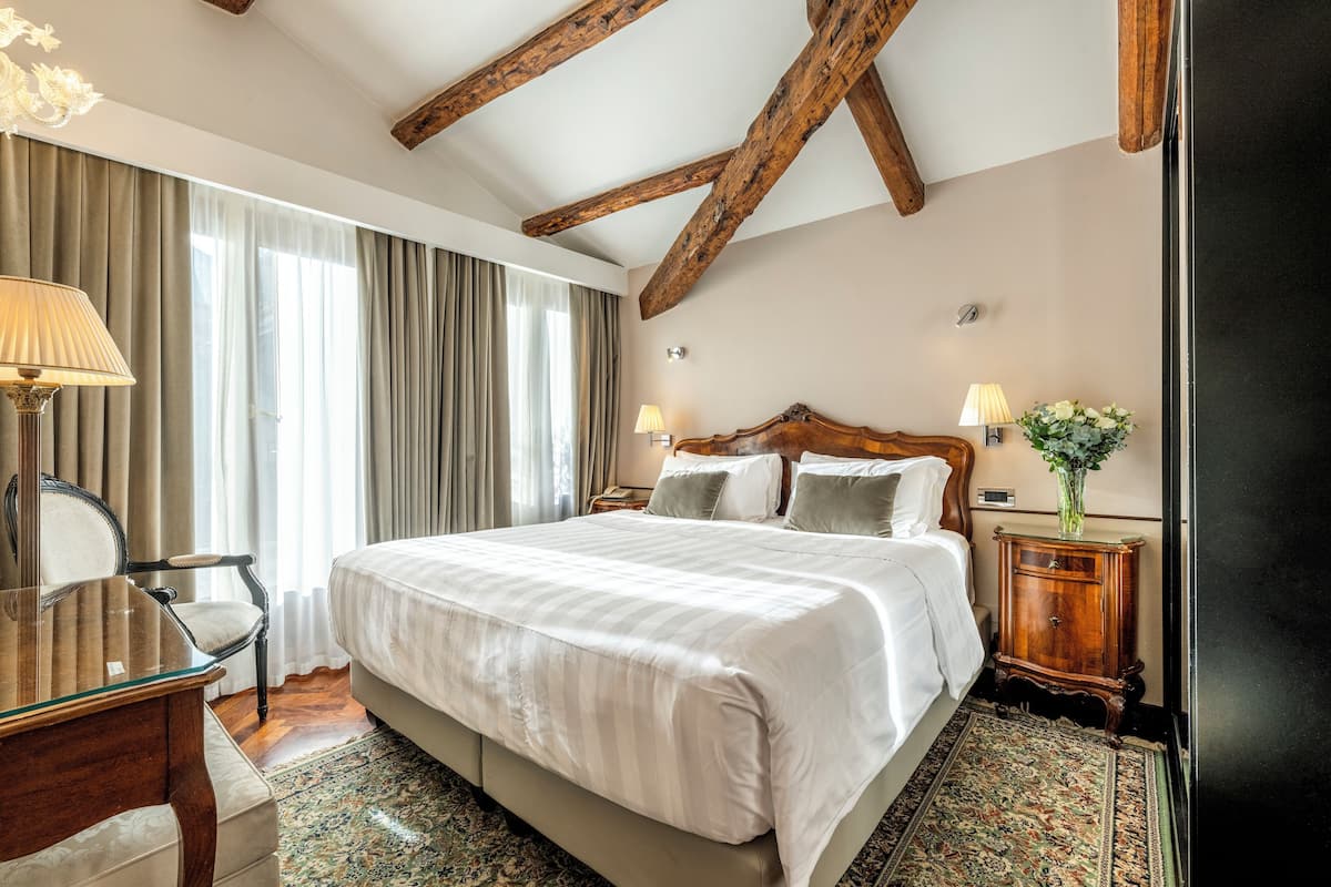 A hotel room with a large bed, wooden beams on the ceiling, a desk with a chair, and a nightstand with a lamp and vase.