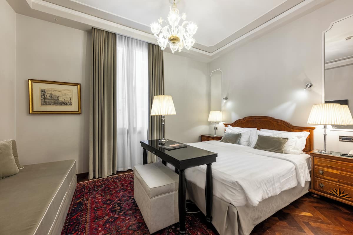 A hotel room with a bed, bedside tables, a desk, a chair, a small table, a bench, a chandelier, a mirror, and a window with curtains.