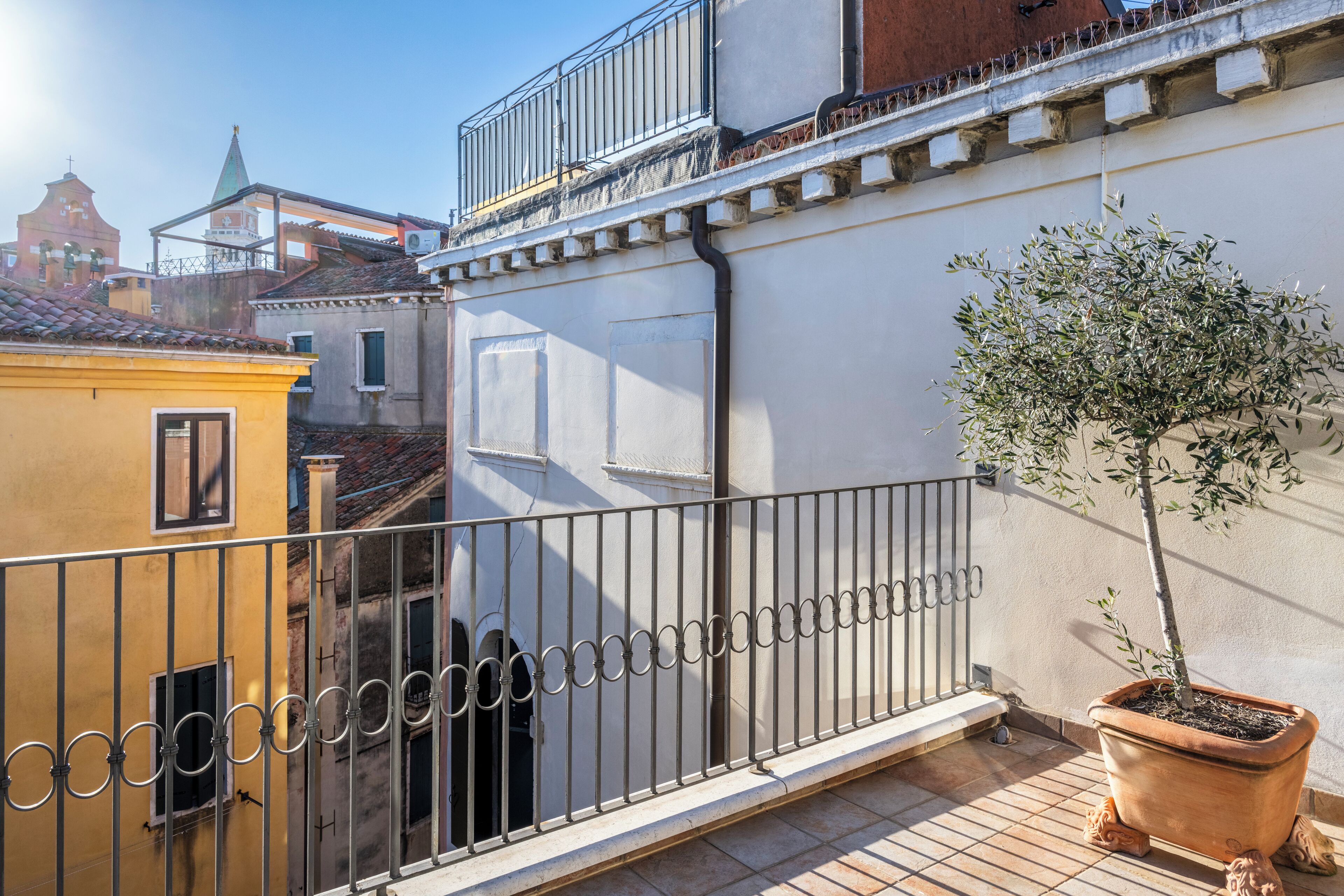 A balcony with a potted tree, a metal railing, and a view of adjacent buildings.