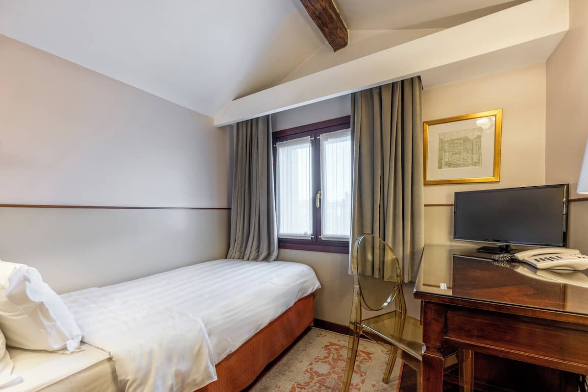A hotel room with a single bed, a desk with a telephone, a chair, a television, and a window with curtains.
