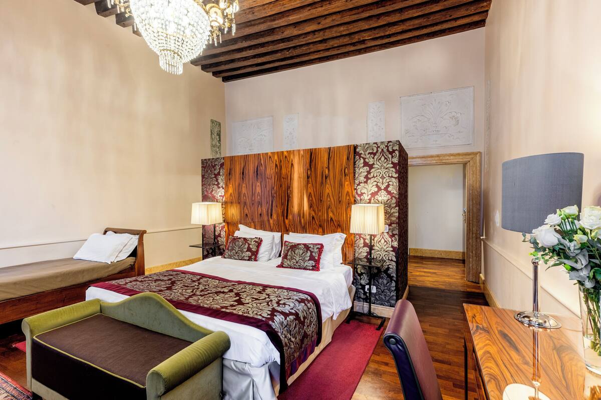 A hotel room with a large bed, two bedside tables with lamps, a wooden headboard, a small sofa, a desk with a lamp, and a vase with flowers.