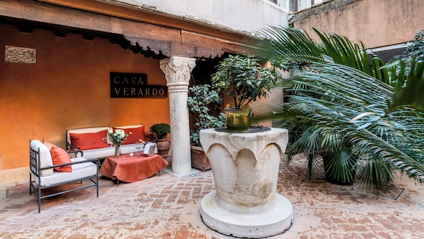 Une cour avec une table en pierre, un canapé avec des coussins rouges et une plante en pot.
