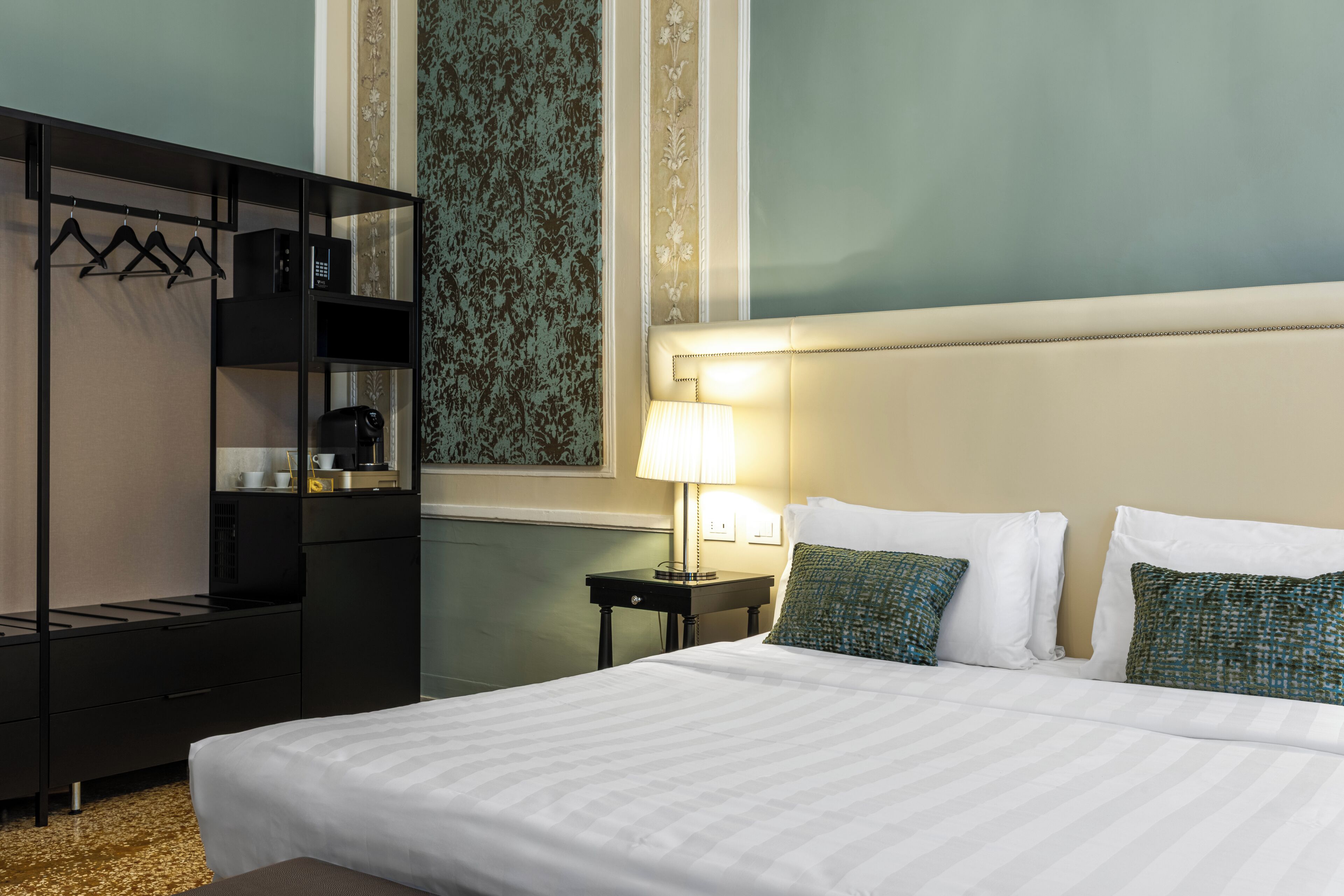 Habitación de hotel con cama, mesita de noche con lámpara, armario y pared con papel tapiz estampado.