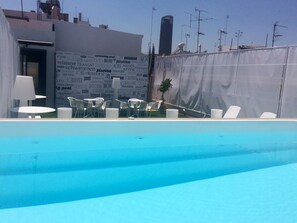 An outdoor pool area with white chairs and tables, a covered seating area, and a large building with text in multiple languages. - Hotel AACR Museo (Seville)