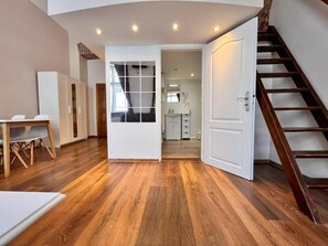 A spacious room with wooden flooring, a dining area with a table and chairs, a staircase, and a view into a kitchen area.