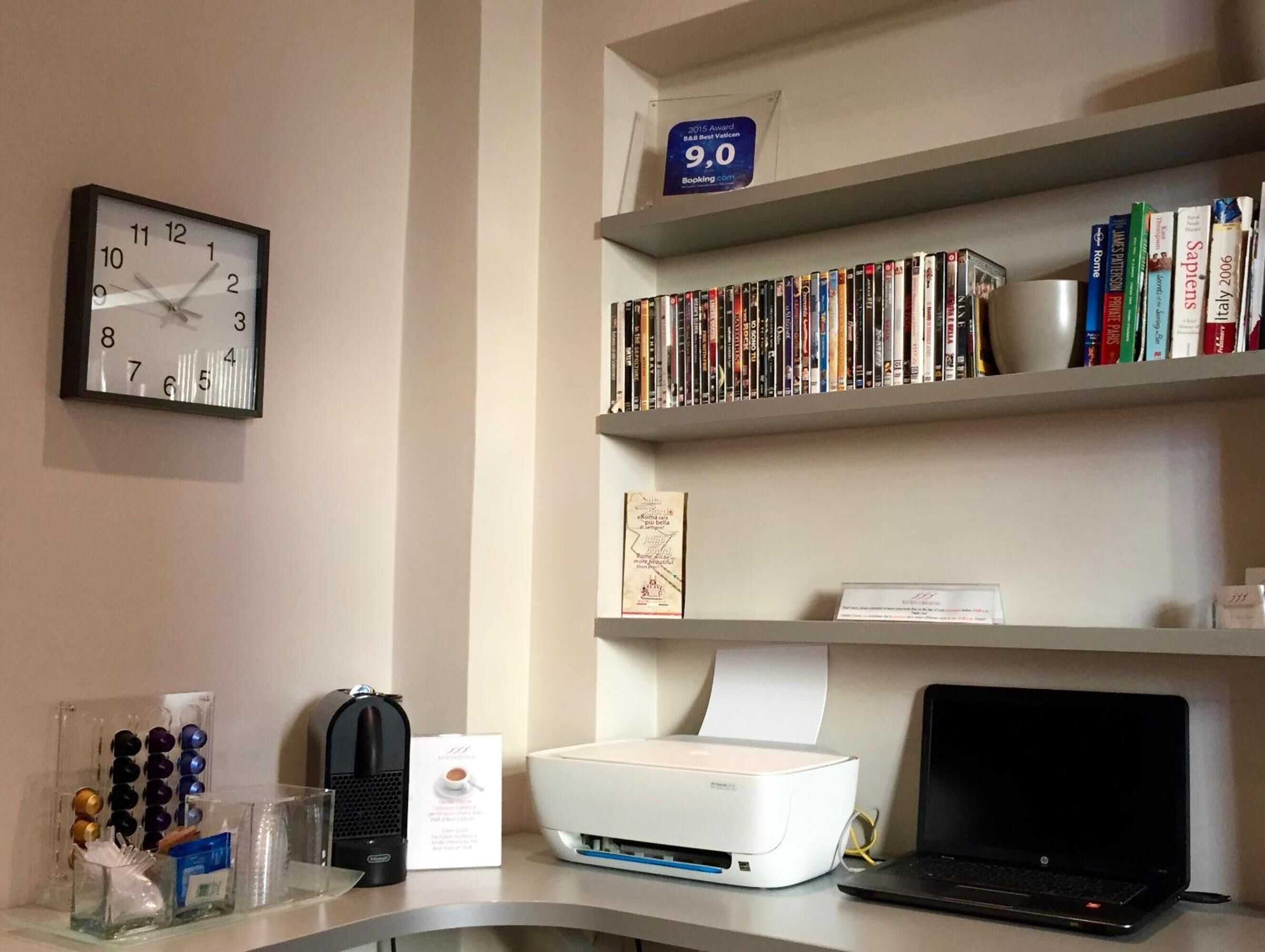 A desk with a clock, a printer, a laptop, and a bookshelf with books.