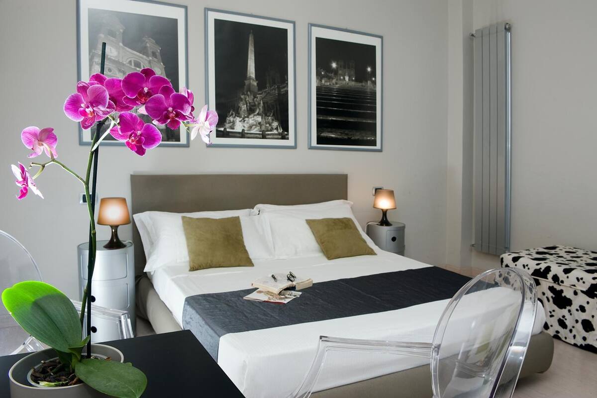 A hotel room with a bed, two bedside tables with lamps, a chair, a desk with a laptop, a potted plant, and framed pictures on the wall.