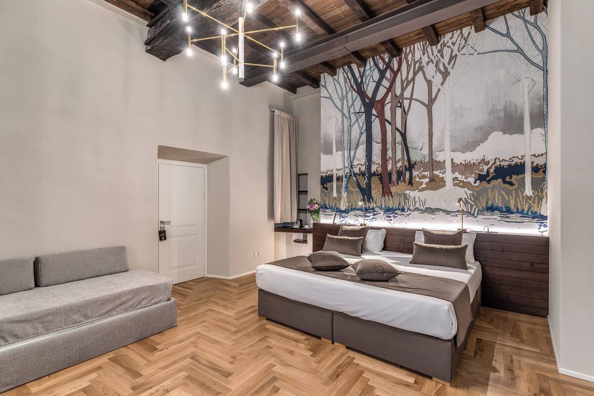 Un dormitorio con una cama grande, un sofá gris, suelo de madera, una lámpara en el techo y un mural en la pared.