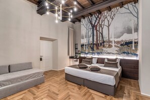 A bedroom with a large bed, a grey sofa, wooden flooring, a ceiling light fixture, and a mural on the wall. - Hotel Abruzzi (Rome)