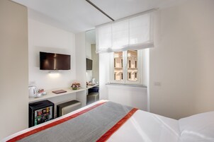 A hotel room with a bed, a TV mounted on the wall, a small kitchenette, and a window with curtains.
