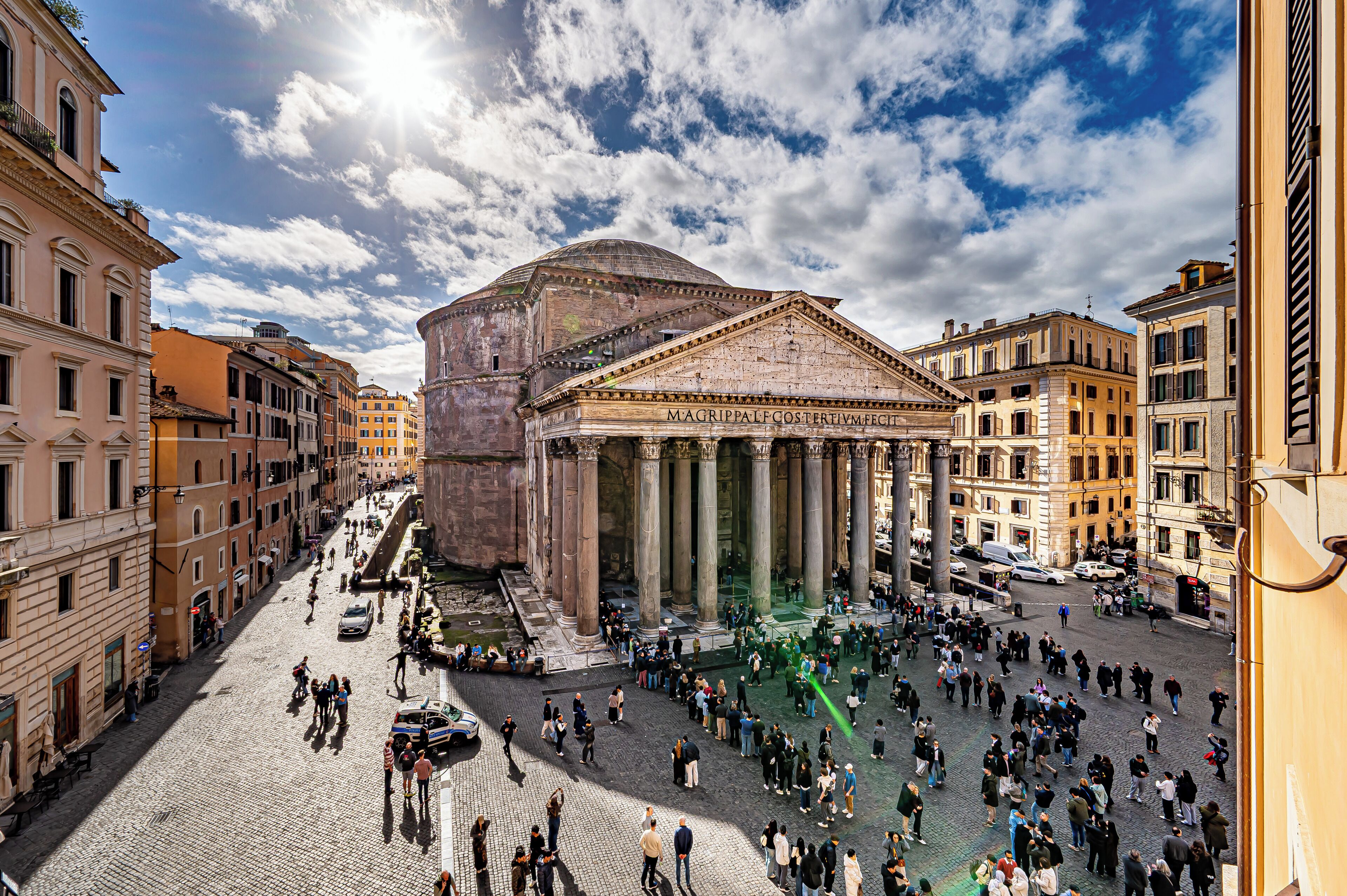 A historic building with classical architecture, including columns and a dome, surrounded by a cobblestone square with pedestrians.