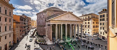 A historic building with classical architecture, including columns and a dome, surrounded by a cobblestone square with pedestrians.