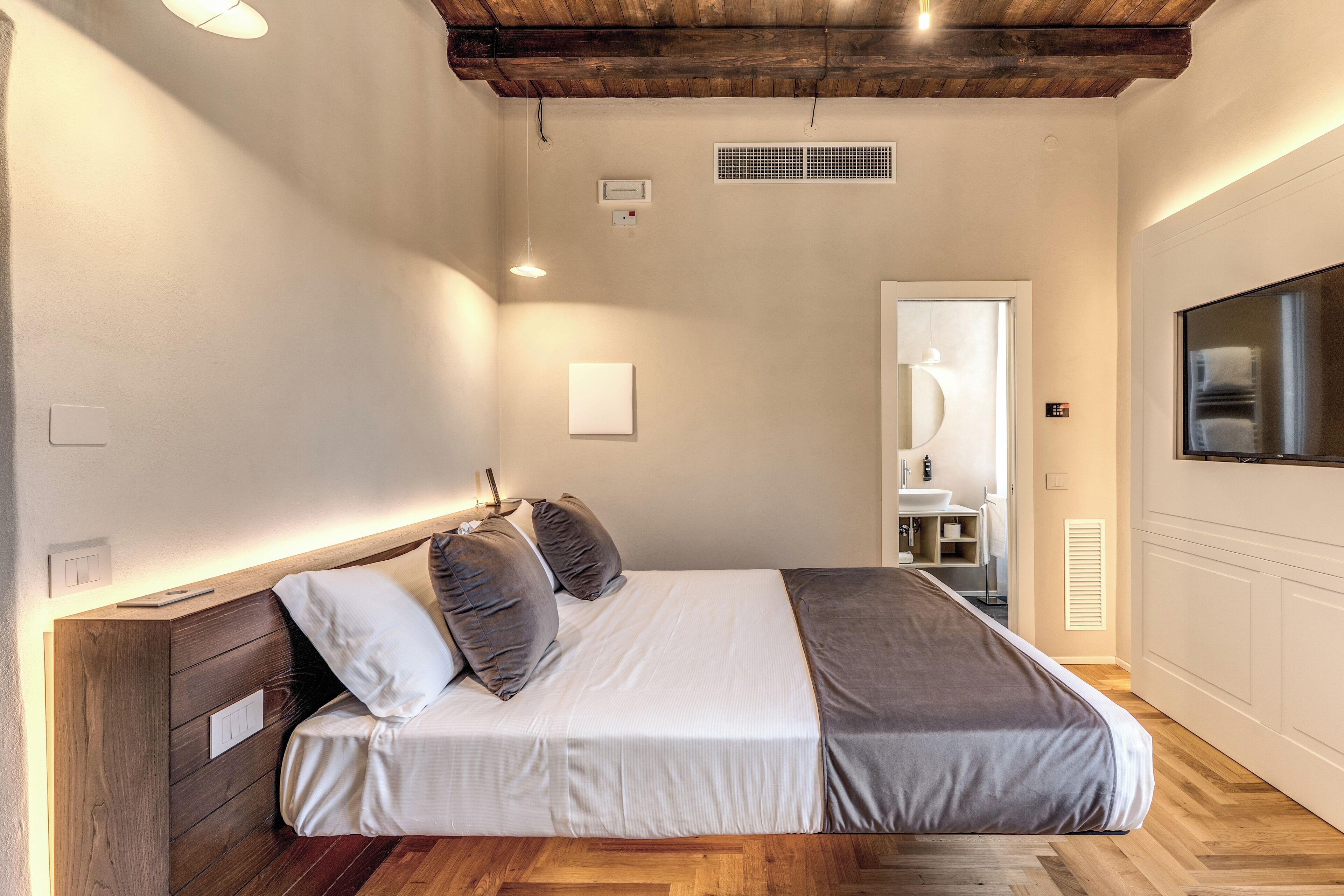 Ein modernes Schlafzimmer mit einem Holz-Kopfteil, einem großen Bett mit grauer Bettwäsche, einem integrierten Fernseher und einem Holzboden.