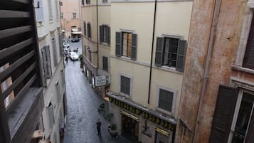 Una estrecha calle adoquinada flanqueada por edificios de varios pisos, algunos con contraventanas, y zonas de estar al aire libre con mesas y sillas.