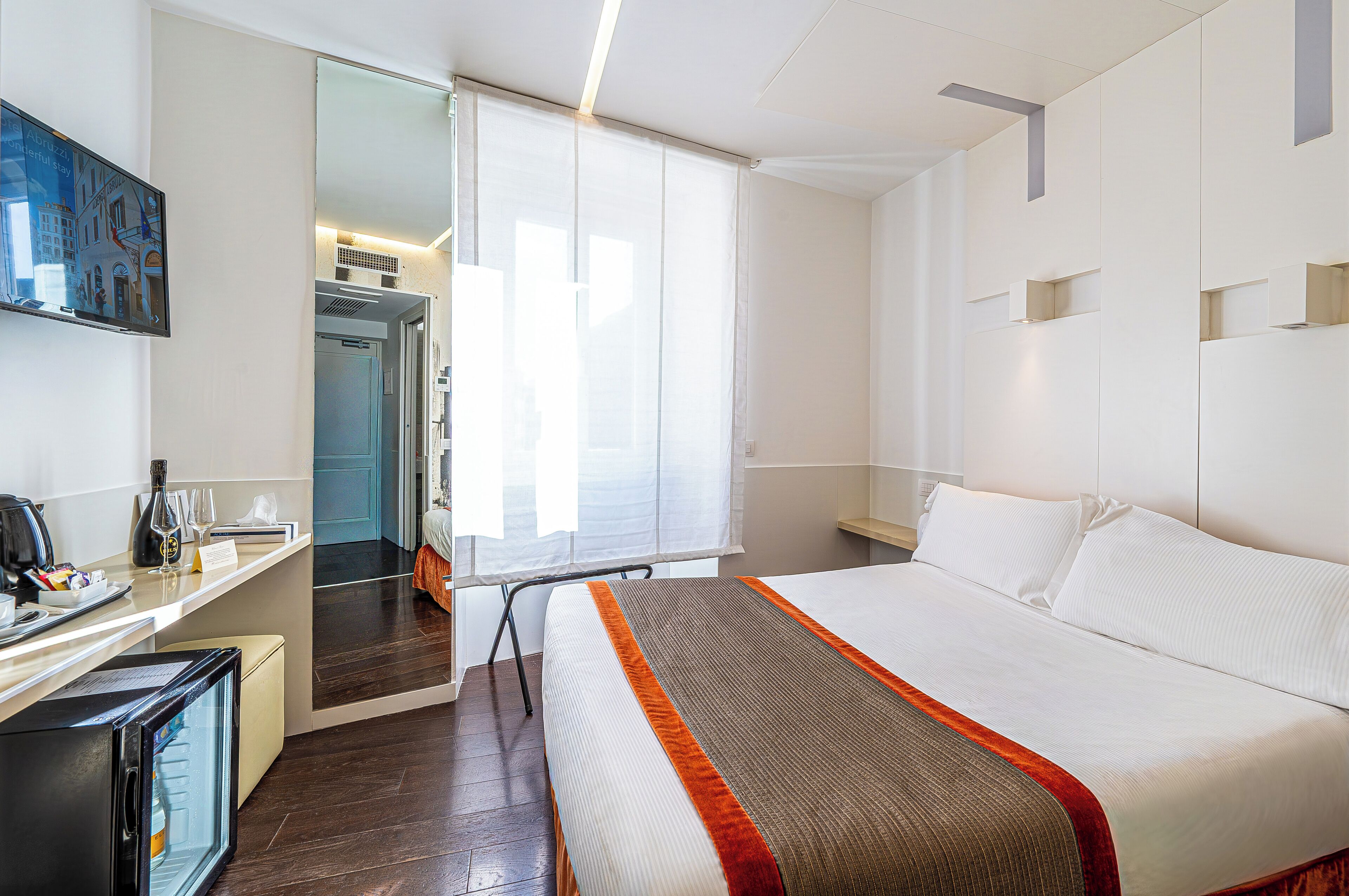 Une chambre d’hôtel équipée d’un lit, d’un bureau avec un ordinateur, d’un mini-réfrigérateur et d’une fenêtre avec des stores.