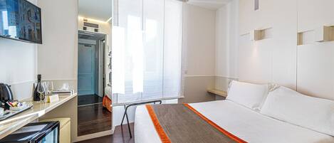 Habitación de hotel con cama, escritorio, computadora, mini refrigerador y ventana con persianas.