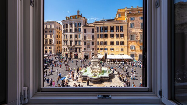 A view through a window of a bustling square with historic buildings and a central fountain.