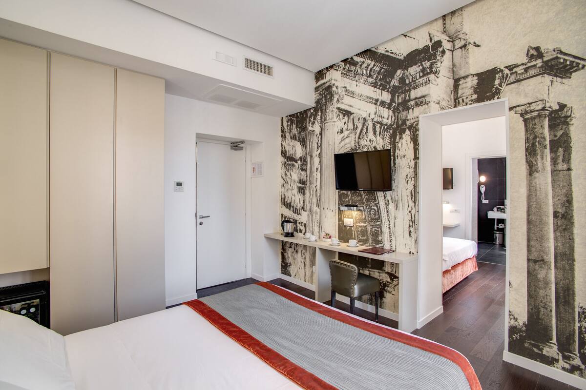 Habitación de hotel con cama, escritorio, televisor y baño visible a través de una puerta abierta.