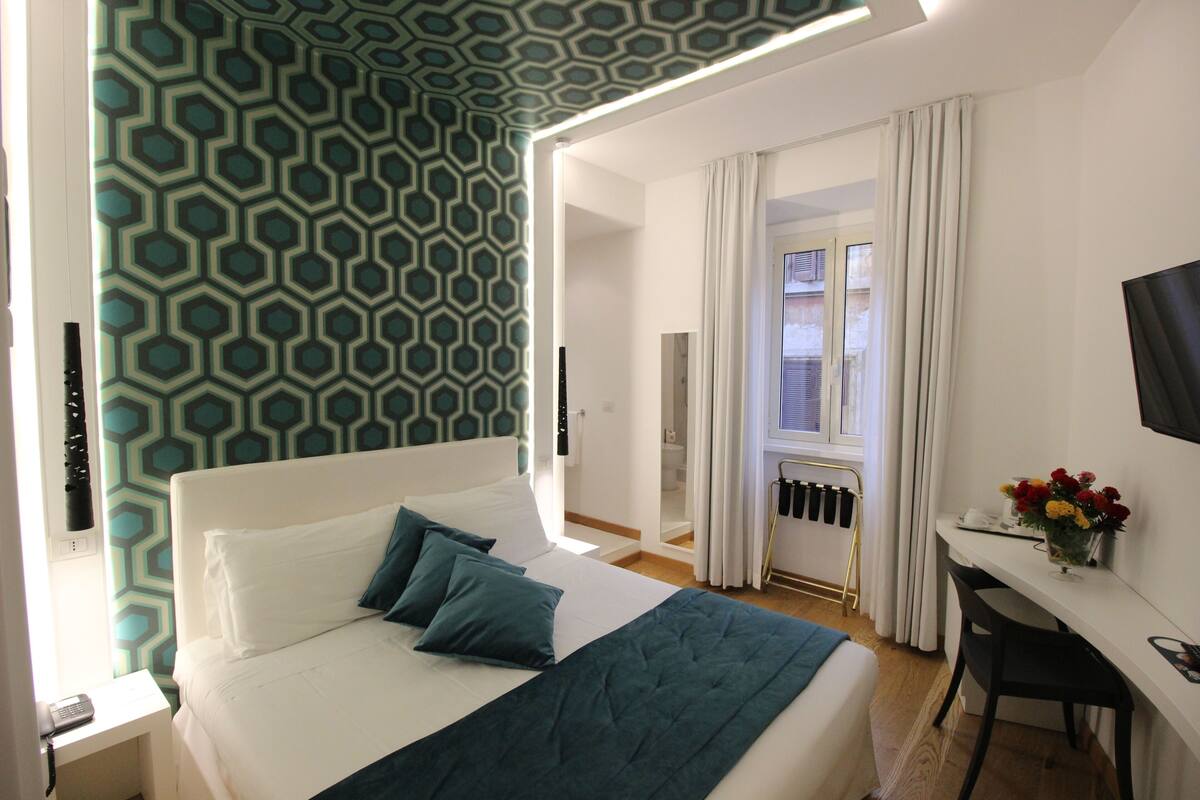 Habitación de hotel con cama, escritorio, silla, televisor y ventana con cortinas.