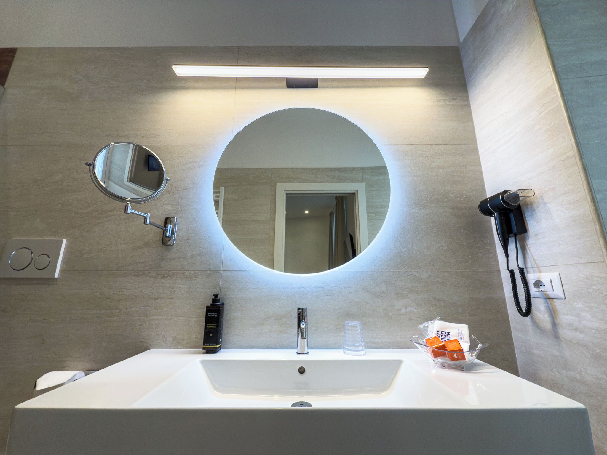 A bathroom with a large oval mirror, a rectangular sink, a handheld phone, and a small bottle of hand soap.