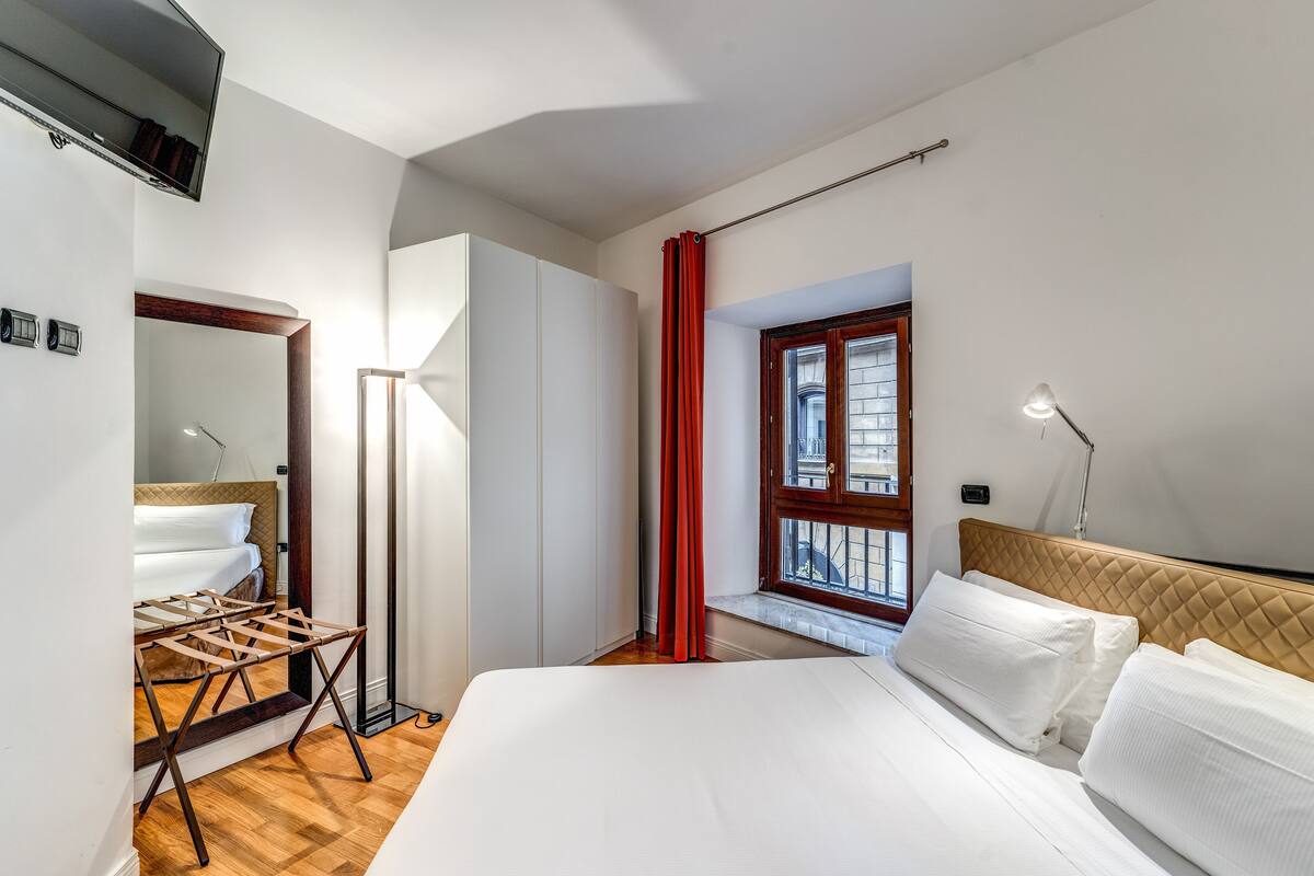 Una habitación de hotel con cama, armario, ventana con cortinas rojas y lámpara de noche.