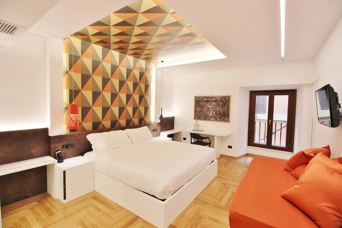 Un dormitorio moderno con techo estampado geométricamente, una cama blanca, un sofá naranja y un escritorio con una silla.