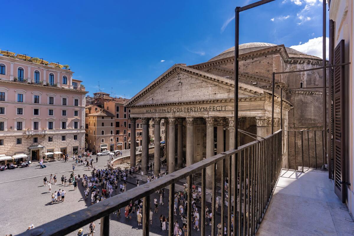 Un edificio histórico con arquitectura clásica, que incluye columnas y frontón, ubicado en una plaza pública concurrida.
